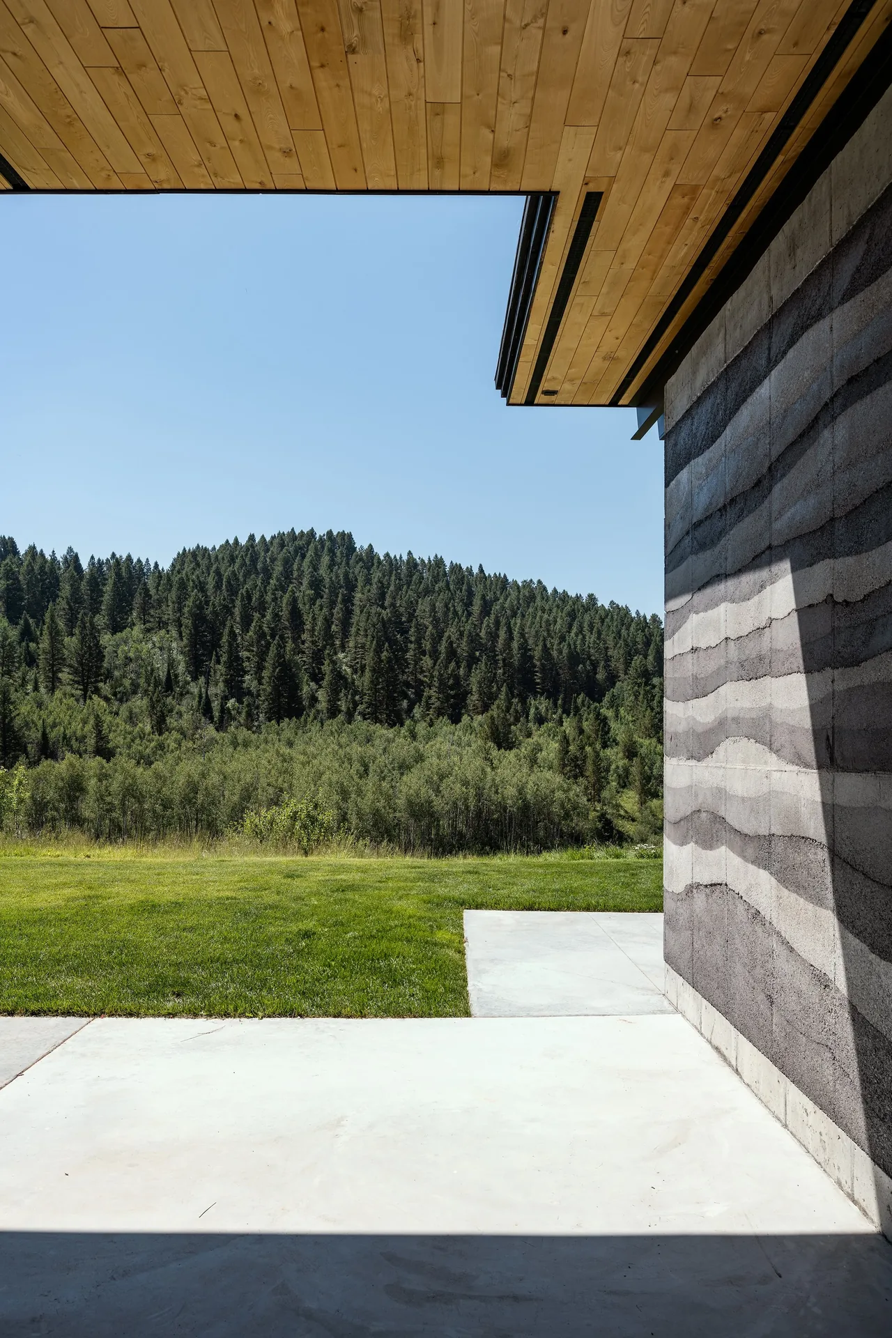 A rammed earth wall is contrasted with a wooden overhang and concrete patio, overlooking green lawns and wooded hills.