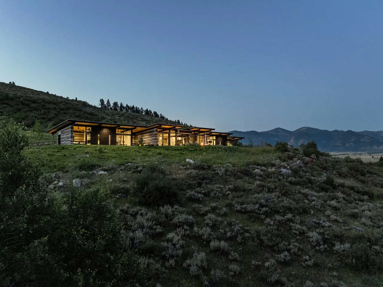 The Freedom Residence features a single-story design with extensive floor-to-ceiling glazing and a flat roof, situated on a hillside at dusk.