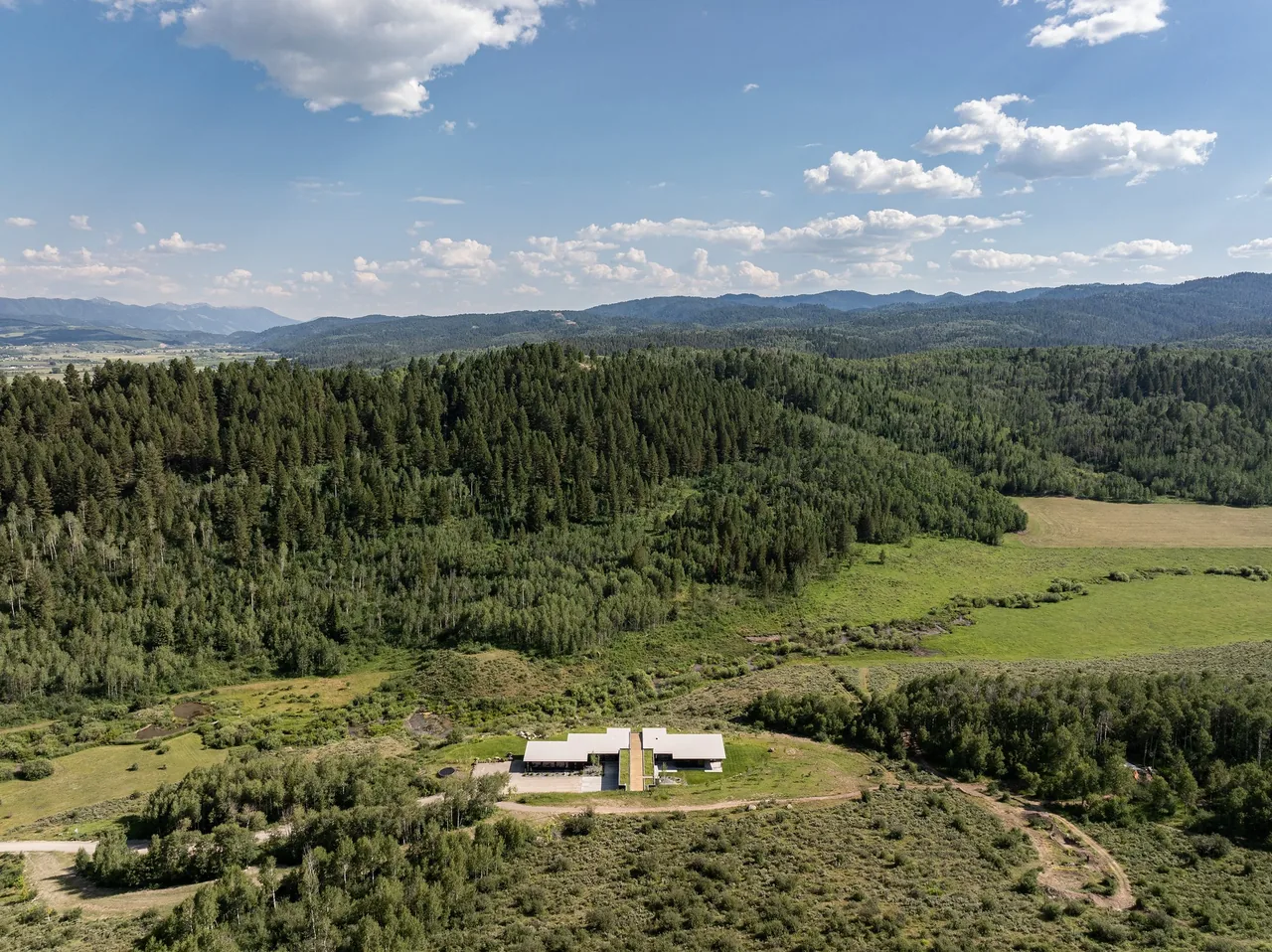 Aerial view shows a modern house with a green roof surrounded by forested hills and open fields.