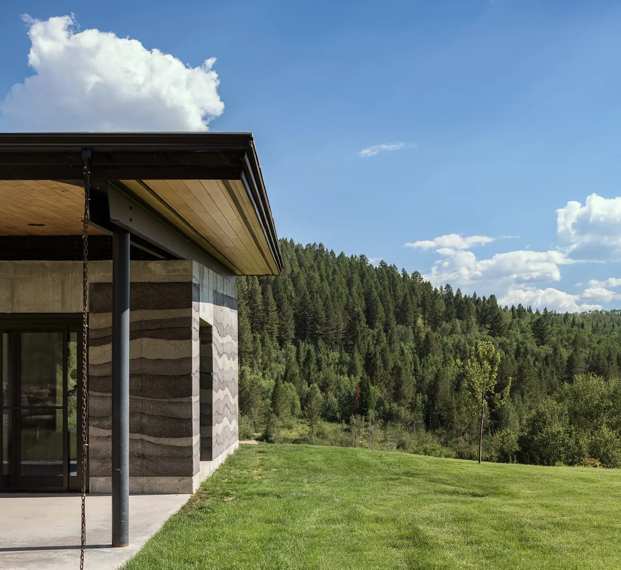 Side view of a modern house showing layered rammed earth walls, concrete base, and an overhanging wooden roof with black steel column.