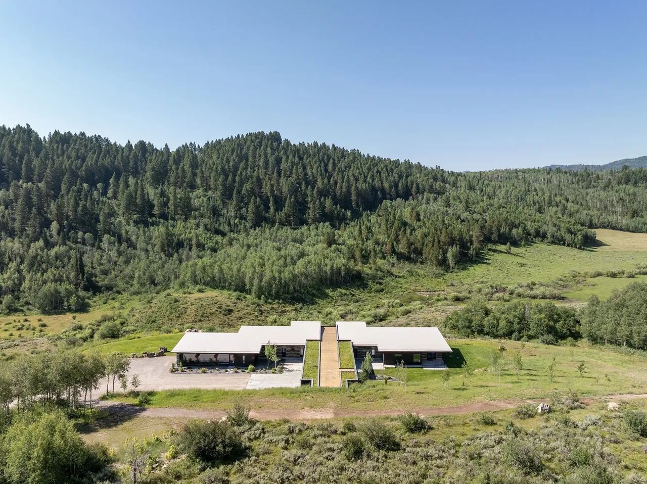 A modern residence with flat roofs and integrated green roof sits in a clearing surrounded by forested hills.