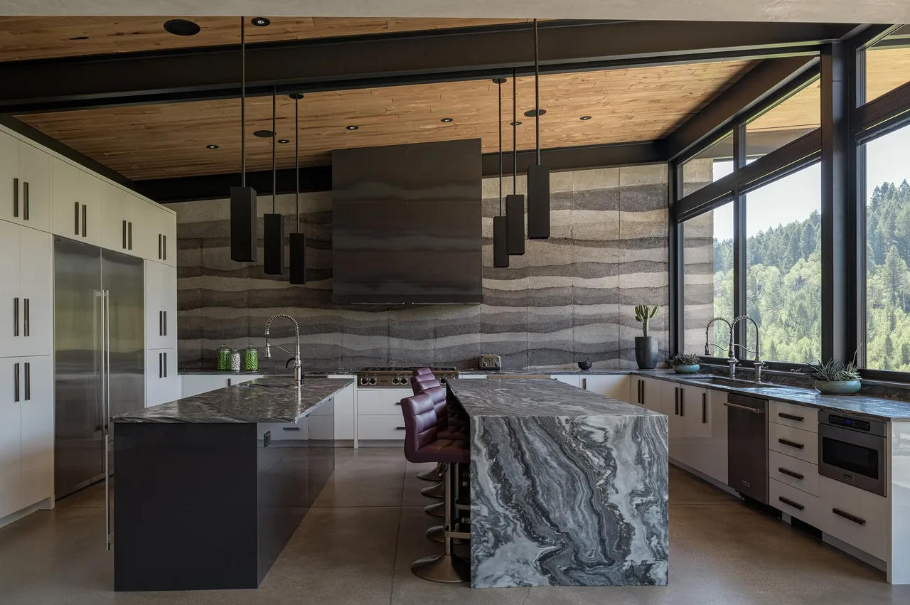 Kitchen featuring two marble islands, large windows with forest views, and a textured layered concrete accent wall.