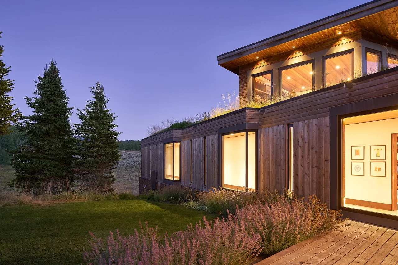 Skyline Ranch Residence features wood siding, black window frames, and large windows with glowing interior light at dusk.