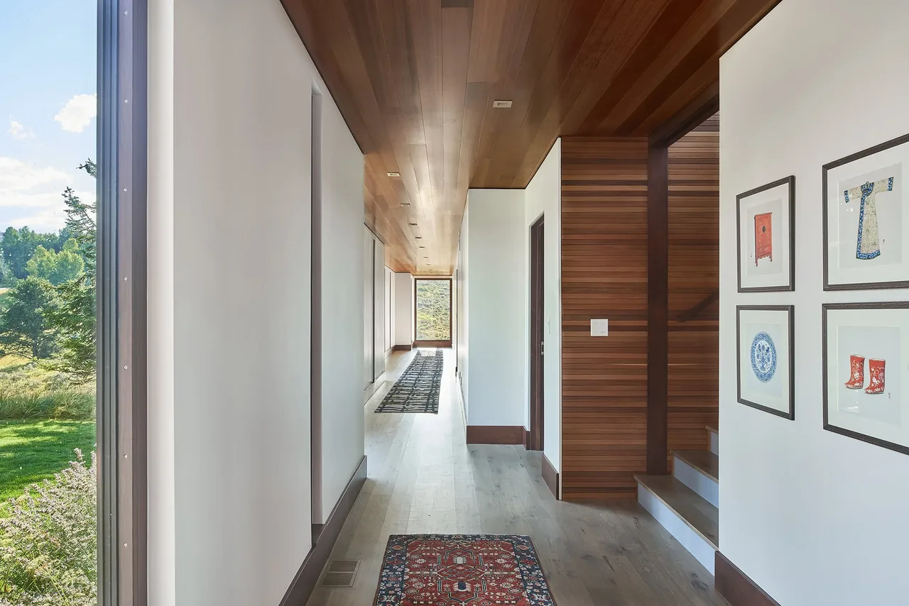 Interior hallway with wooden ceiling and stair walls alongside white walls and rugs leading to a window with an outdoor view.