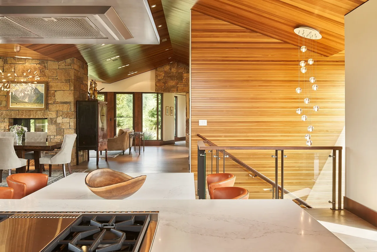 Open interior featuring a kitchen with gas cooktop, wood-paneled stair wall, stone fireplace, and dining area.