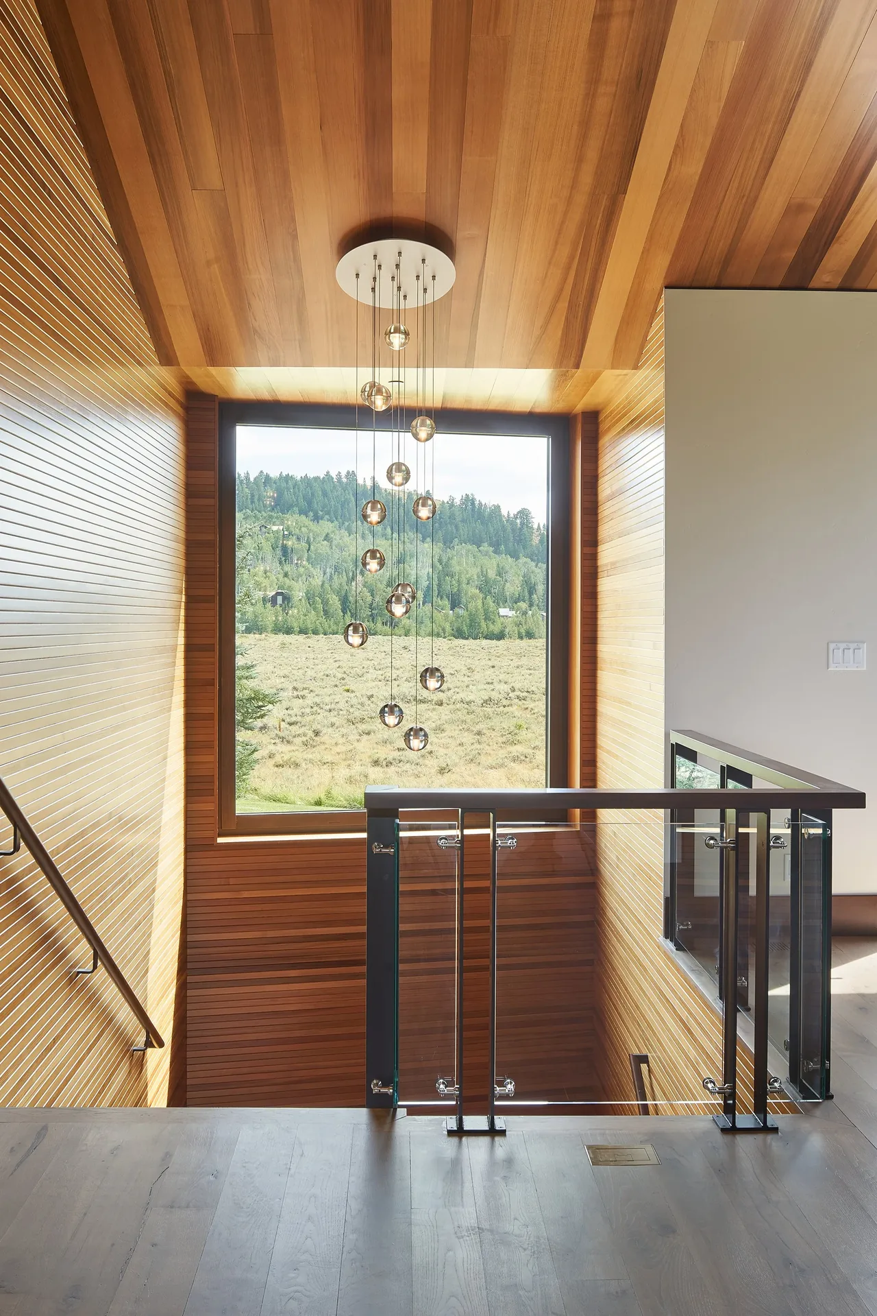Wood-paneled stairwell with modern glass railing and a large window framing a view of forested hills.