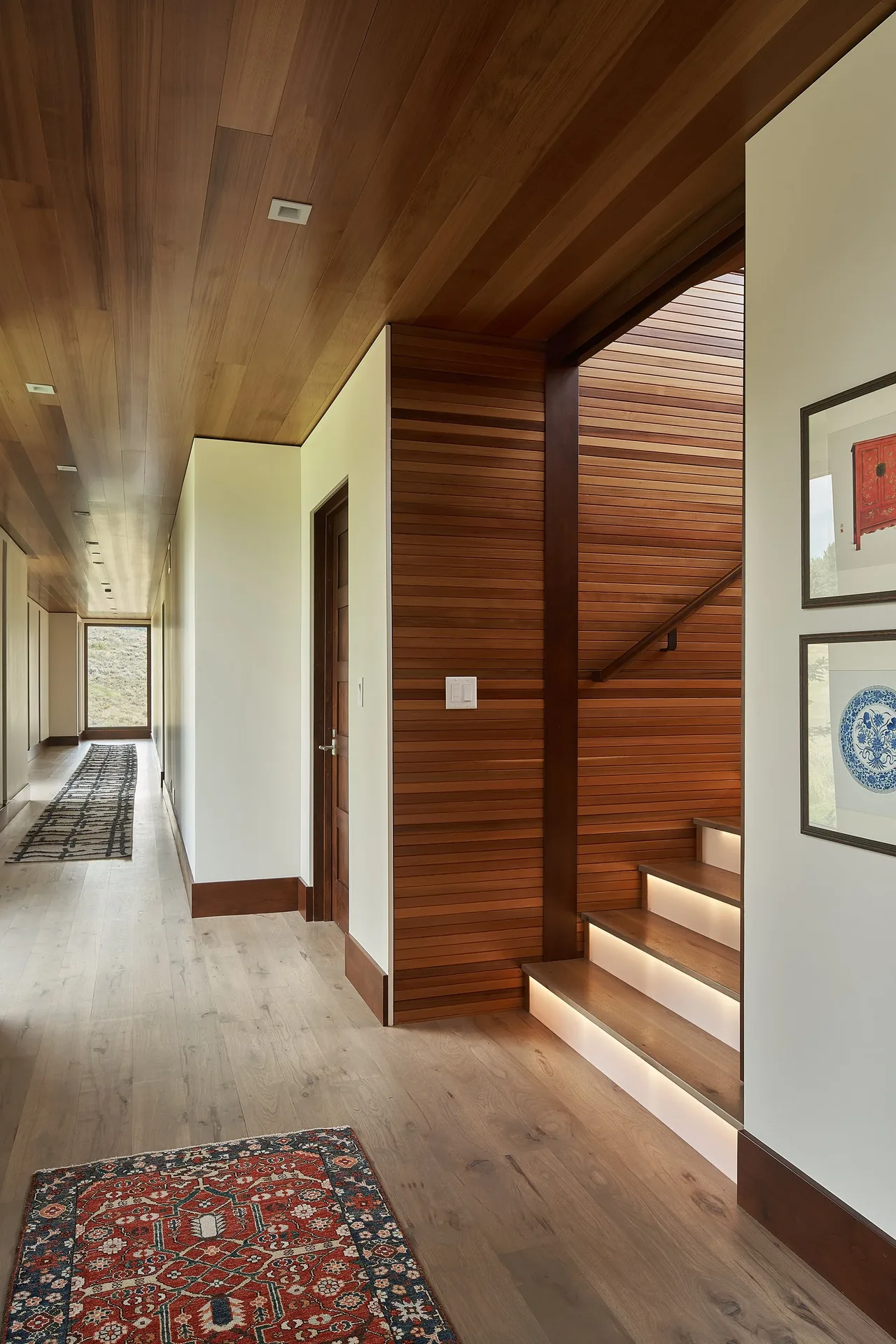 Long hallway features wood ceiling and floor with a wood-paneled staircase illuminated by recessed lighting.