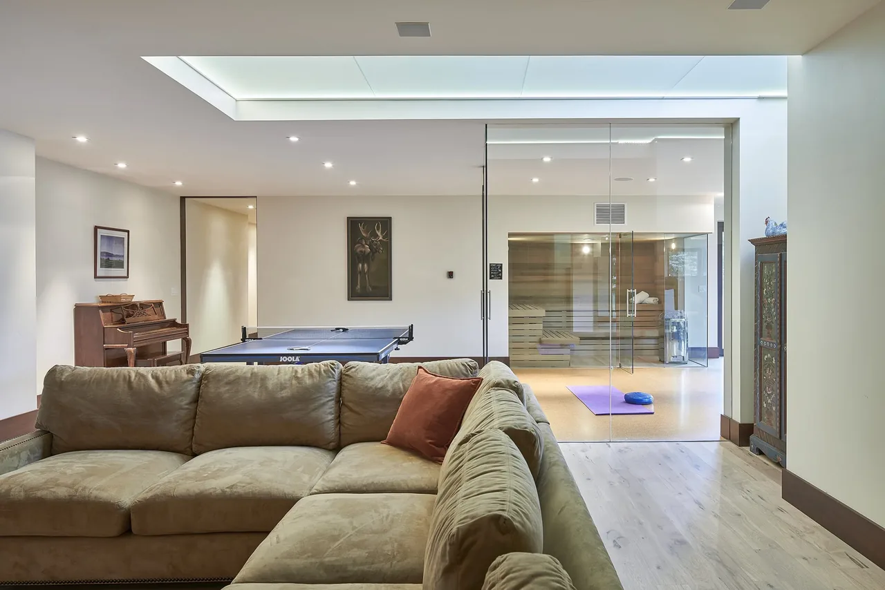 Living room with sectional sofa, piano, ping pong table, and glass-enclosed sauna space visible through a large window.