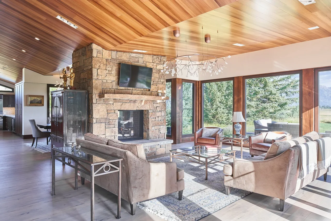 Skyline Ranch Residence living area features a stone fireplace, wood-paneled ceiling, and expansive wood-framed windows overlooking greenery.