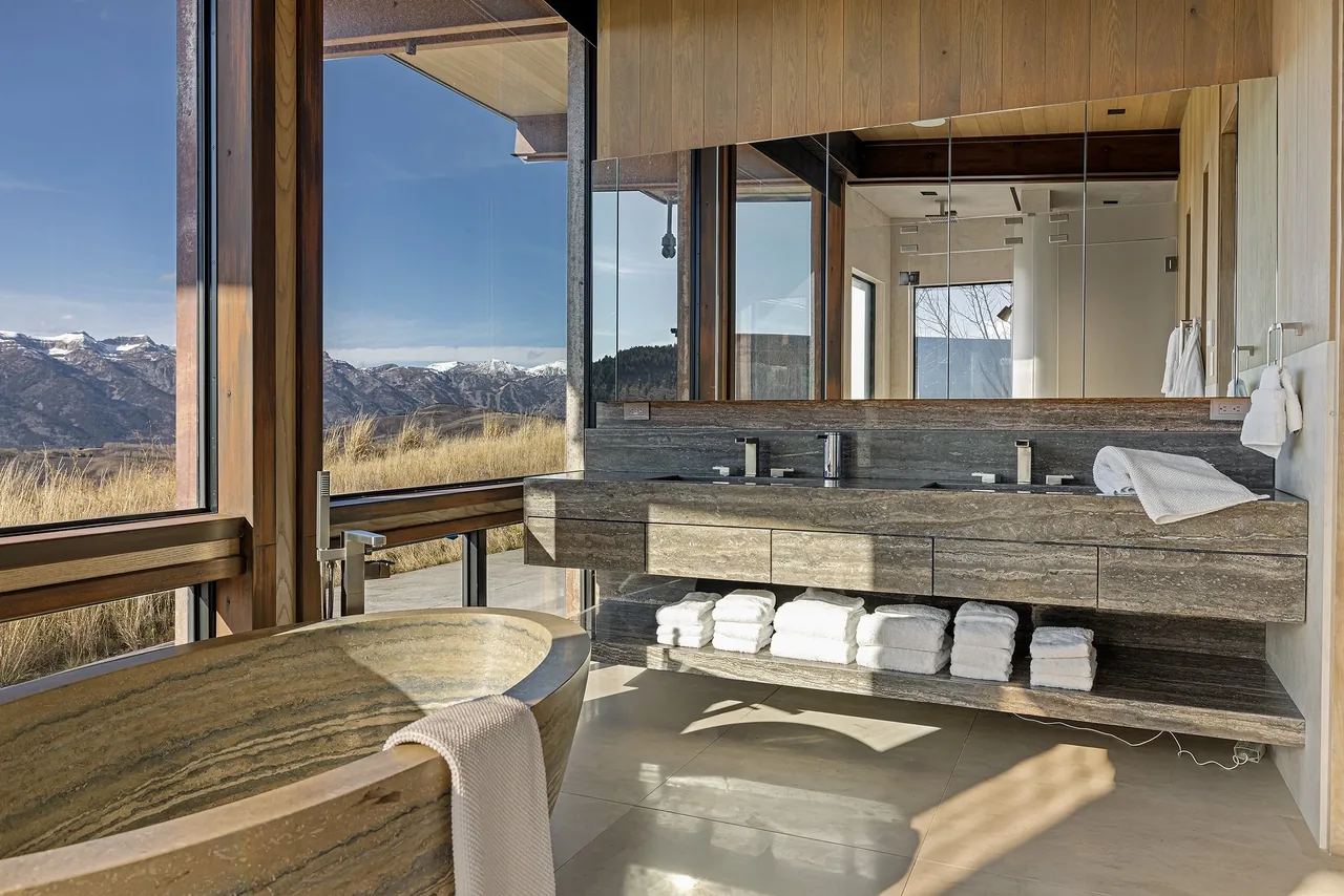 Bathroom featuring a stone bathtub and double vanity with towel storage beneath, illuminated by natural light from expansive windows.
