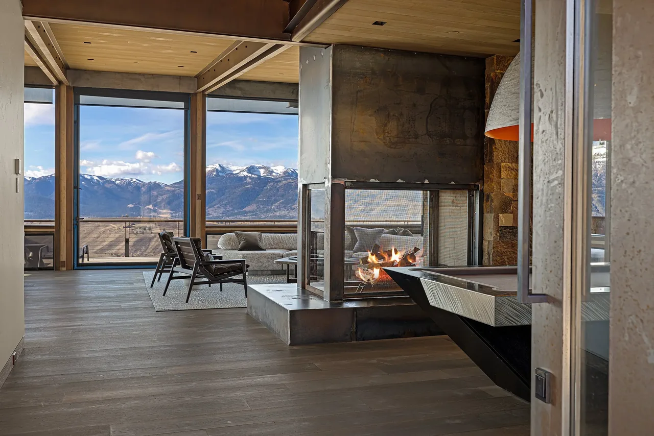 Living room with floor-to-ceiling glazing and a three-sided fireplace overlooking mountains.