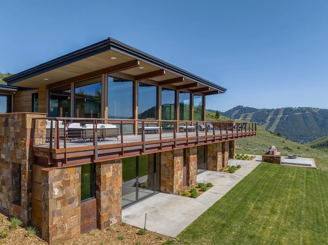 Modern residence with stone facade and expansive upper-level terrace overlooking a landscaped lawn and mountain view.