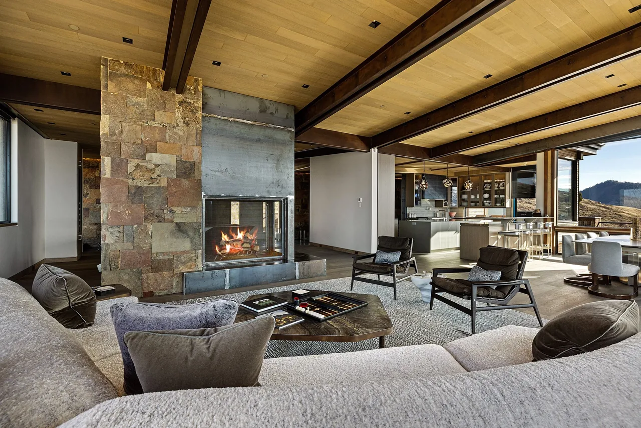 Living room and kitchen space with a double-sided fireplace, exposed wood beams on the ceiling, and floor-to-ceiling glazing.