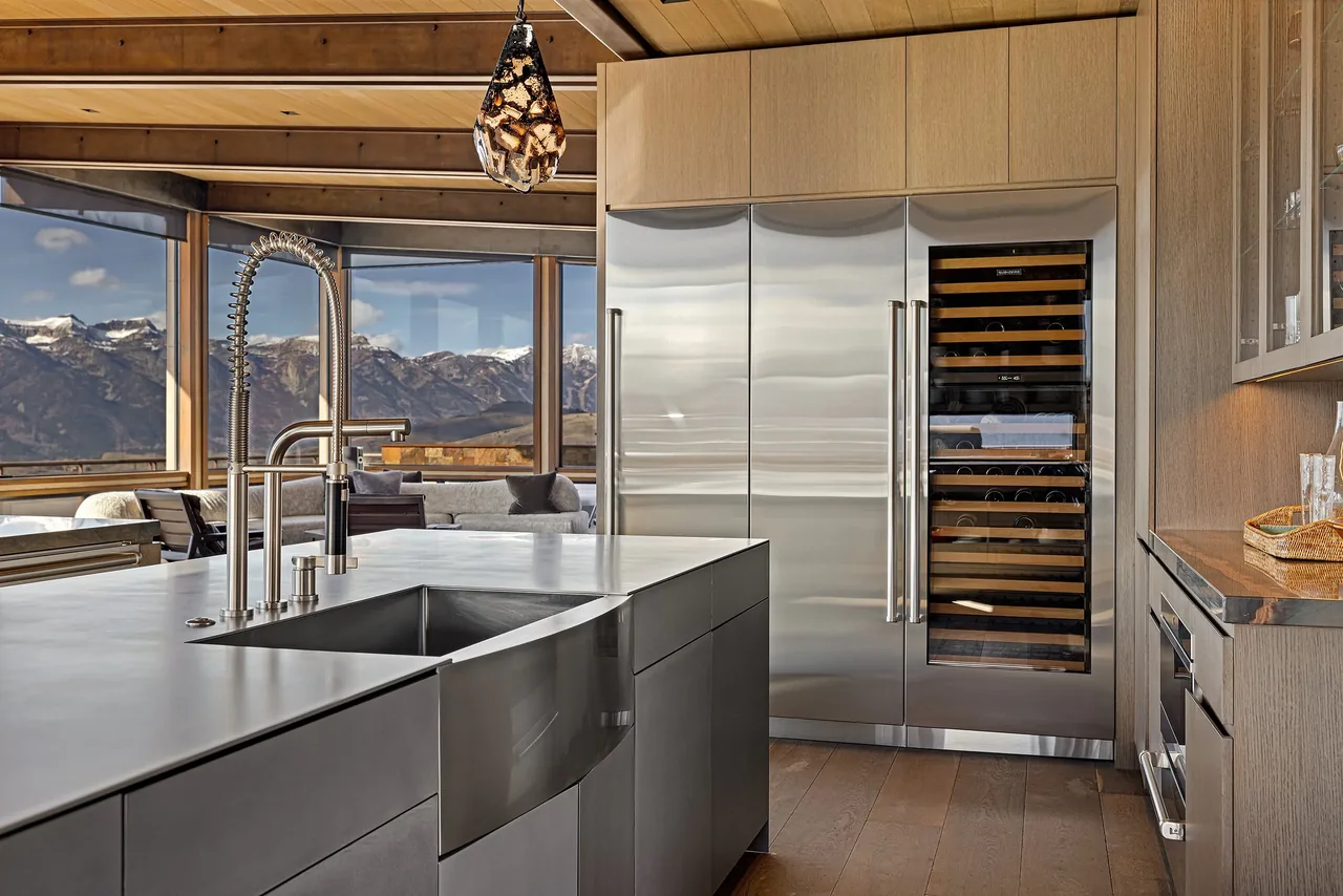 Kitchen featuring stainless steel island with sink and built-in wine refrigerator against wood-paneled cabinets and large windows with mountain views.