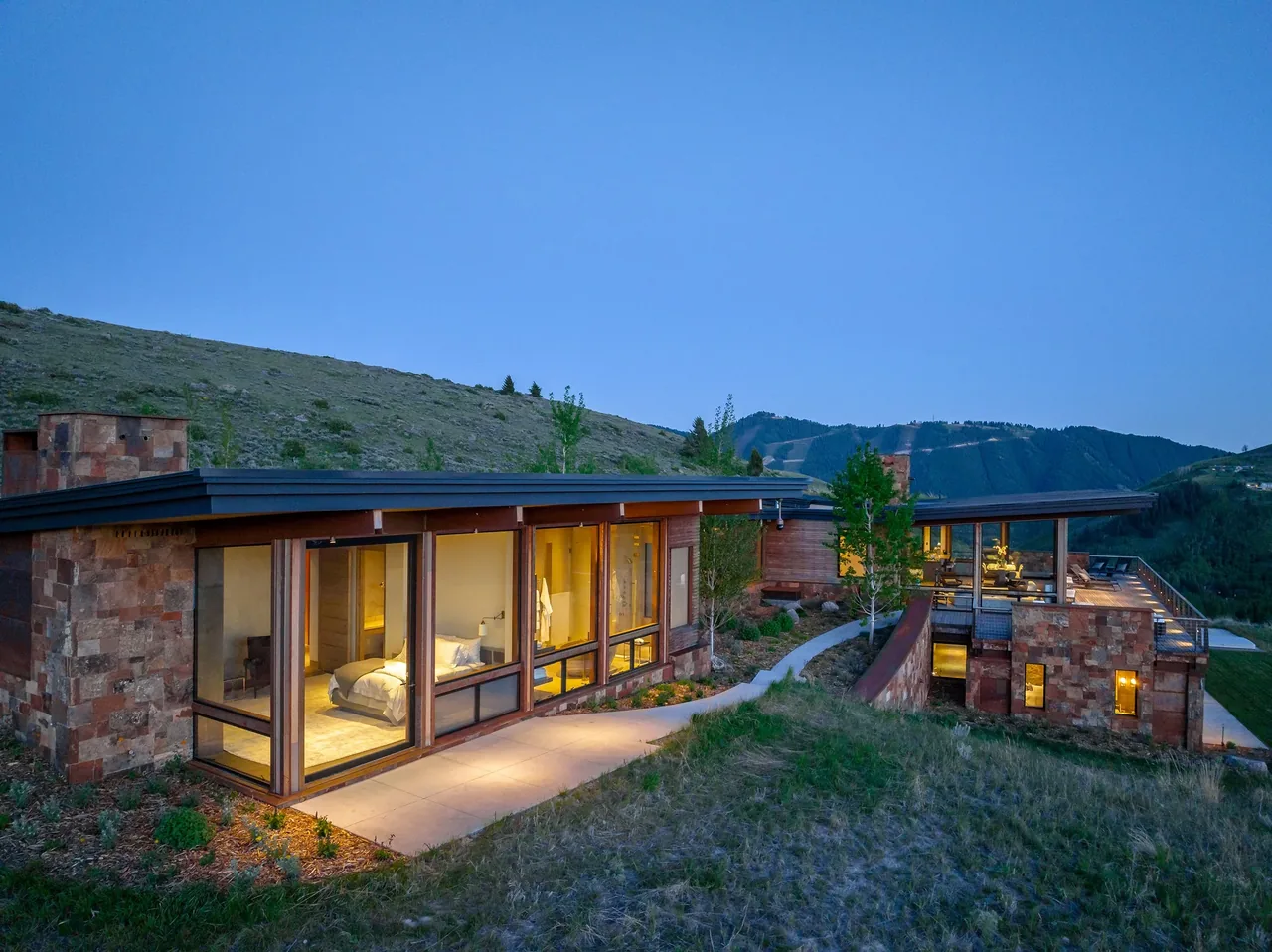 Ridge 52 Residence features stone and glass construction with flat roofs on a hillside setting at dusk.