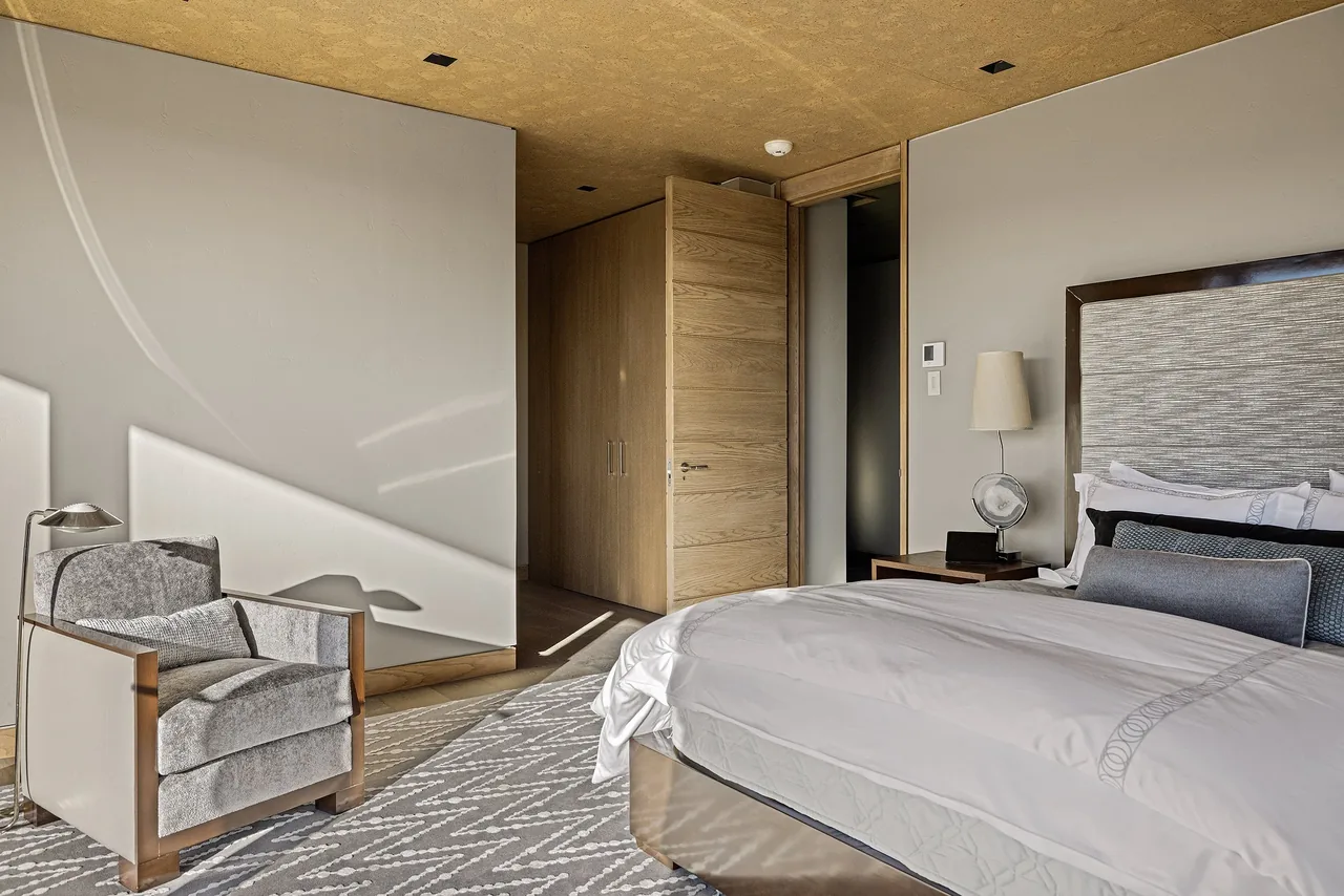 A bedroom featuring a bed with textured headboard, gray armchair, wooden door, and geometric carpet.