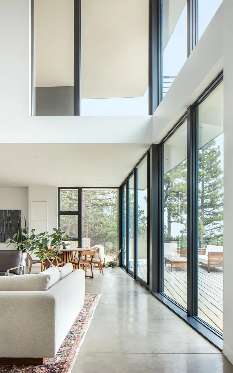 Open living and dining space with black window frames and large windows facing the outdoor deck and forest view.