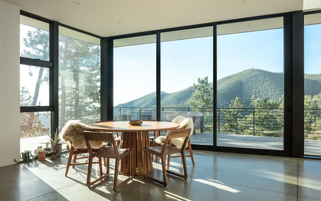 Dining space with wooden furniture and floor-to-ceiling glazing overlooks forested mountains.