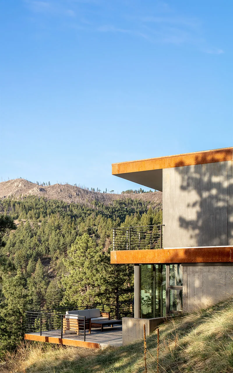 Puma Run house features a concrete and rusted metal exterior with a balcony and outdoor seating overlooking forested hills.