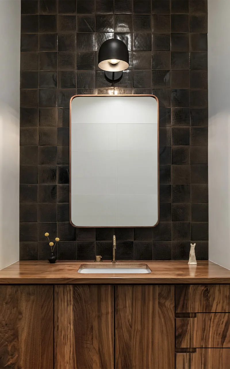 Modern bathroom vanity with walnut cabinetry, dark ceramic tile wall, and minimalist black wall sconce lighting.