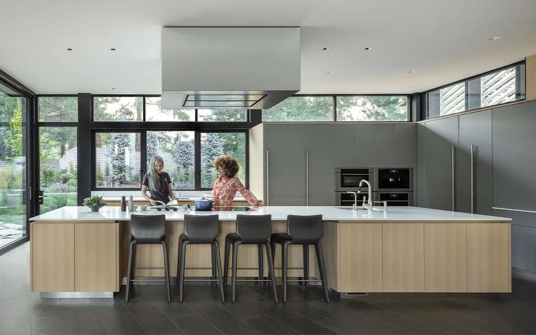 A modern kitchen interior features a large wooden island with bar seating and built-in grey cabinetry under ample natural light.