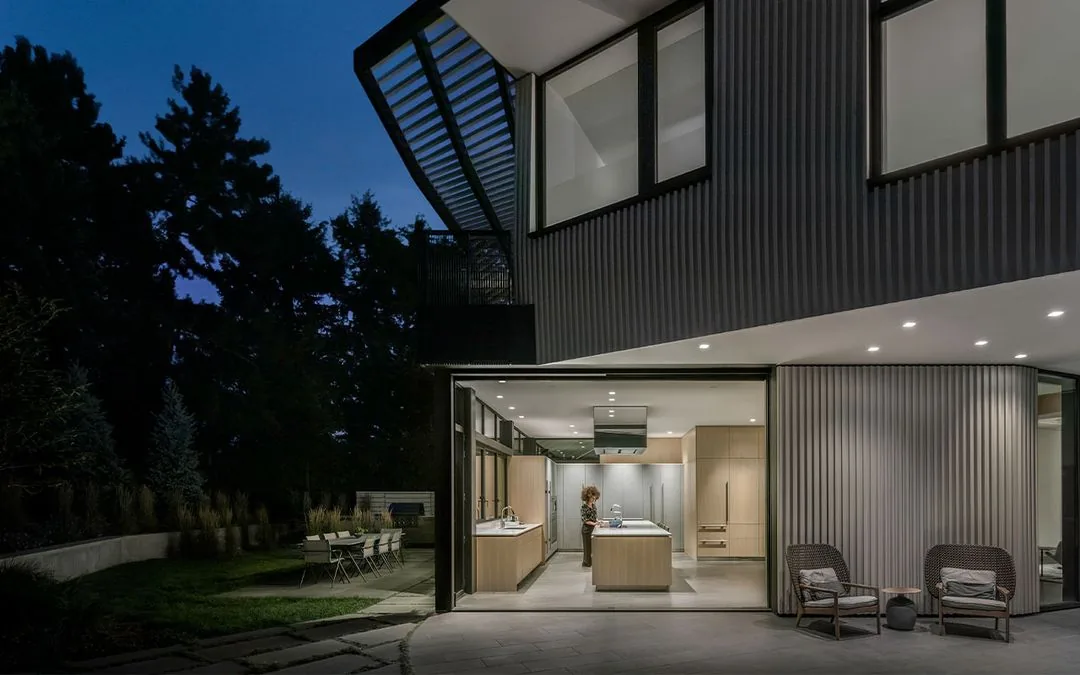 Exterior night view of a modern house with an open kitchen visible through large glass walls and outdoor seating area.