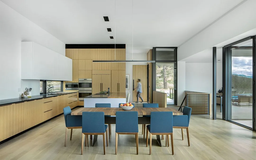 Open-plan kitchen and dining area featuring wood cabinetry, blue chairs, and floor-to-ceiling glass doors.