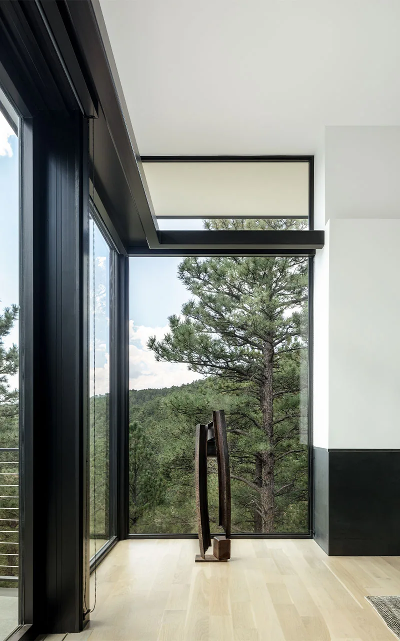 Interior corner with floor-to-ceiling corner windows featuring black frames and a sculptural metal artwork on light wood flooring.