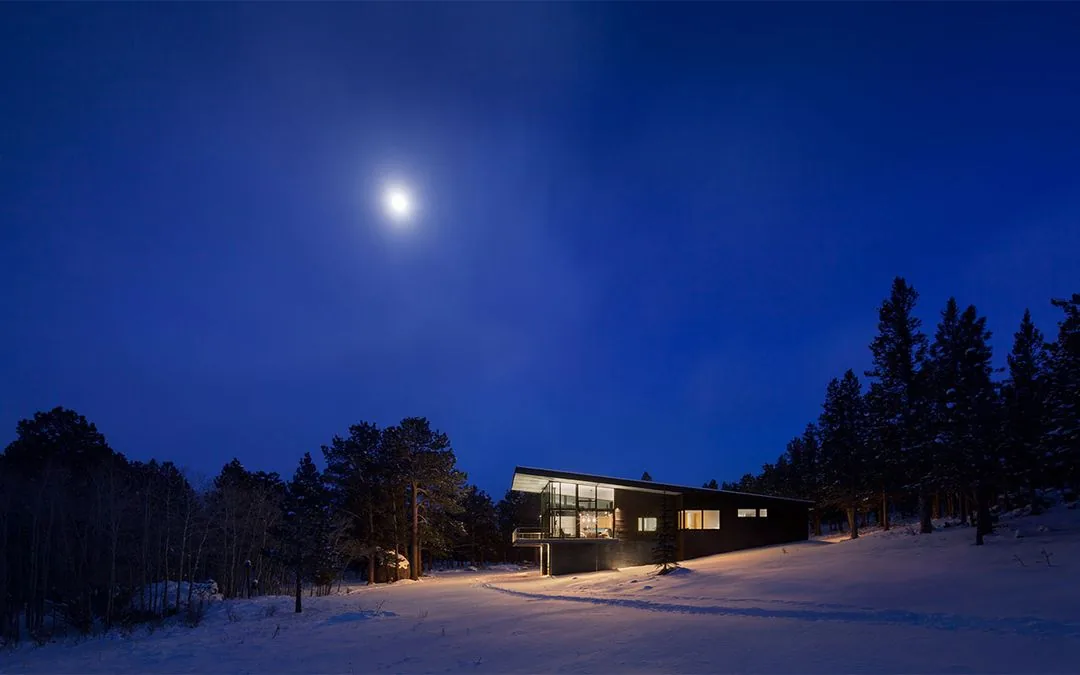 Modern house illuminated at night with large windows and snow-covered landscape under moonlight.