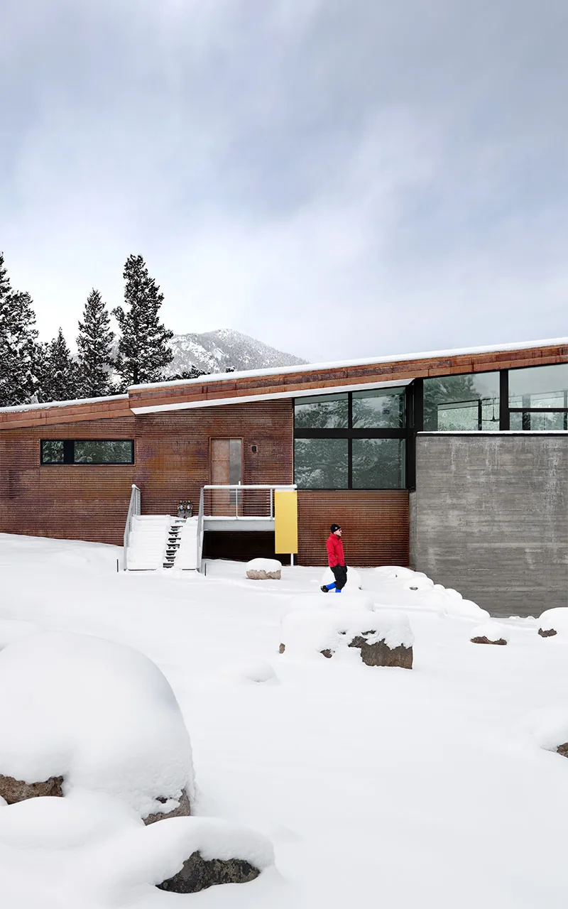 Snow-covered landscape with modern house featuring rust-colored cladding and large windows.