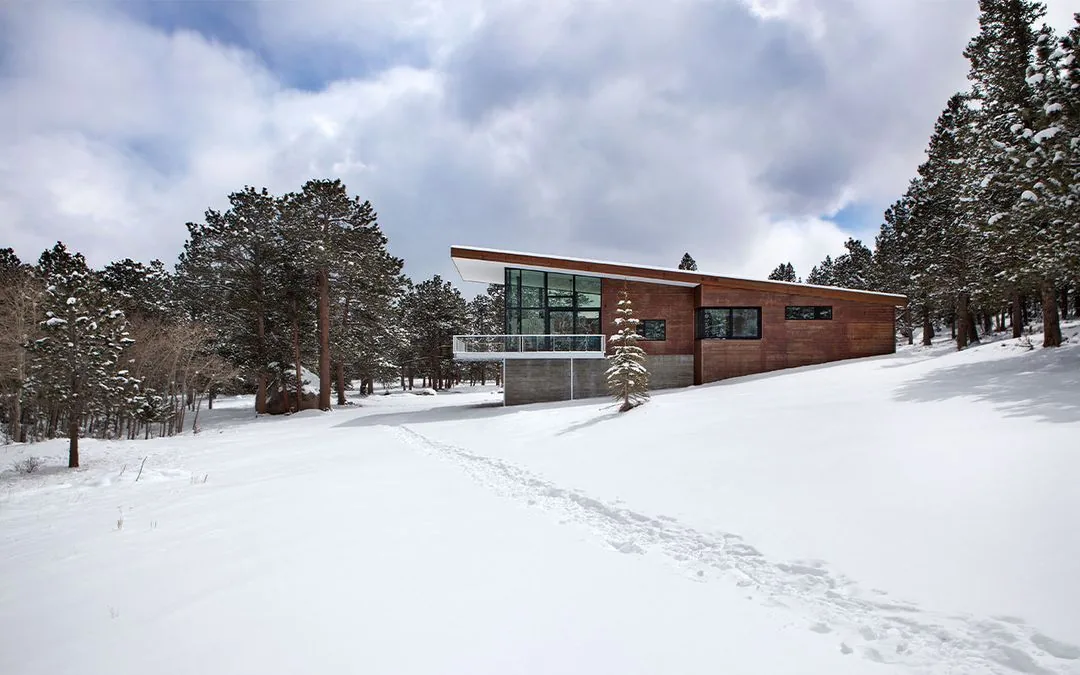 Exterior front view of a modern lodge-style house with wood siding, concrete foundation, and large windows in a snowy landscape.