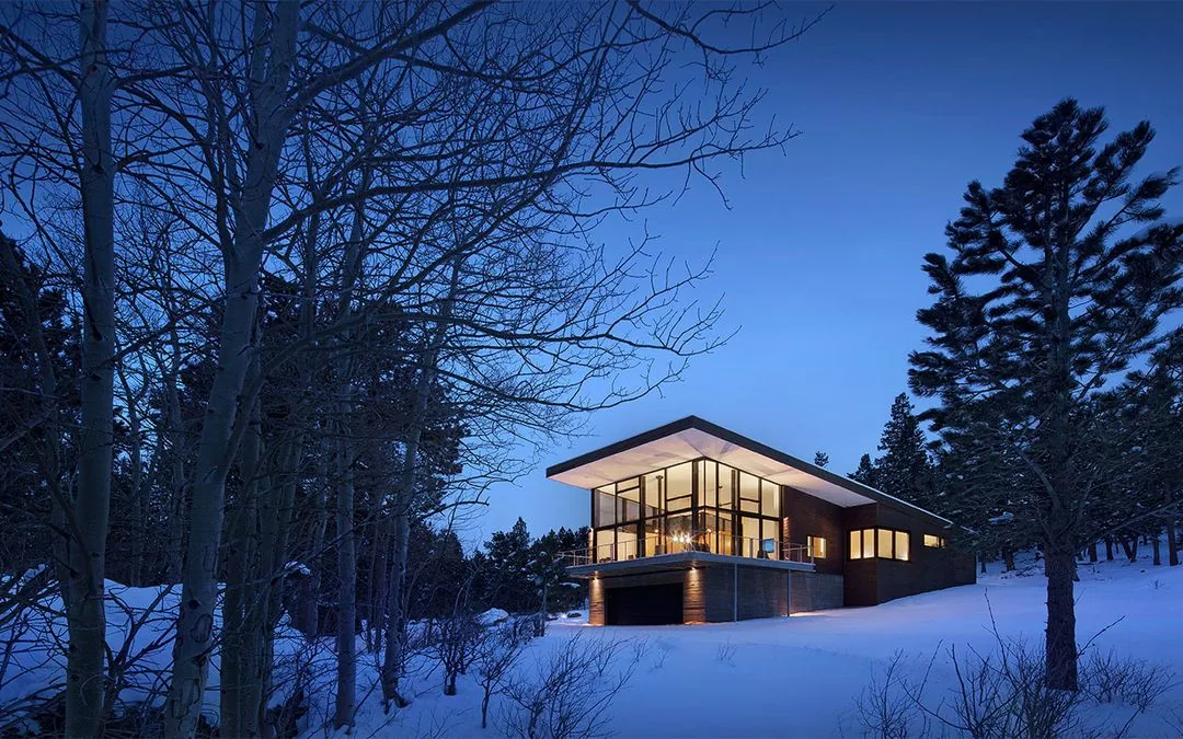Dusk view of a modern house with glass walls and cantilevered roof surrounded by snow-covered trees.