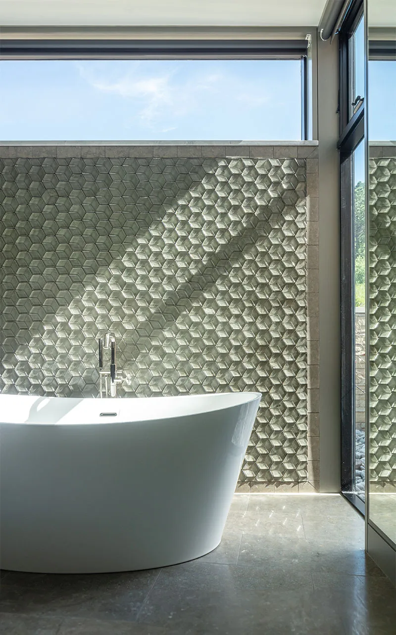 A bathroom with a freestanding tub and a textured hexagonal tile accent wall illuminated by natural light from a high window and glass door.