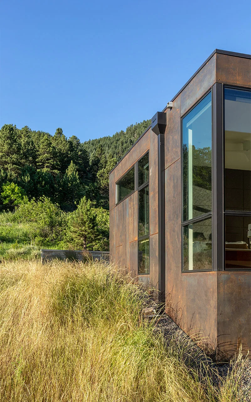 Side elevation of House 306 showing weathered steel cladding and large windows overlooking native grasses and hillside trees.
