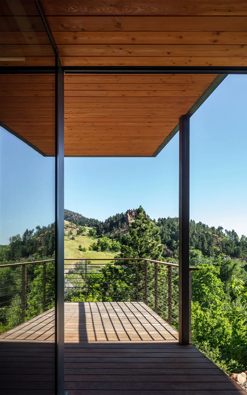 View of a covered balcony with wood ceiling and decking, metal railing, and glass wall reflecting the surrounding green hillside landscape.