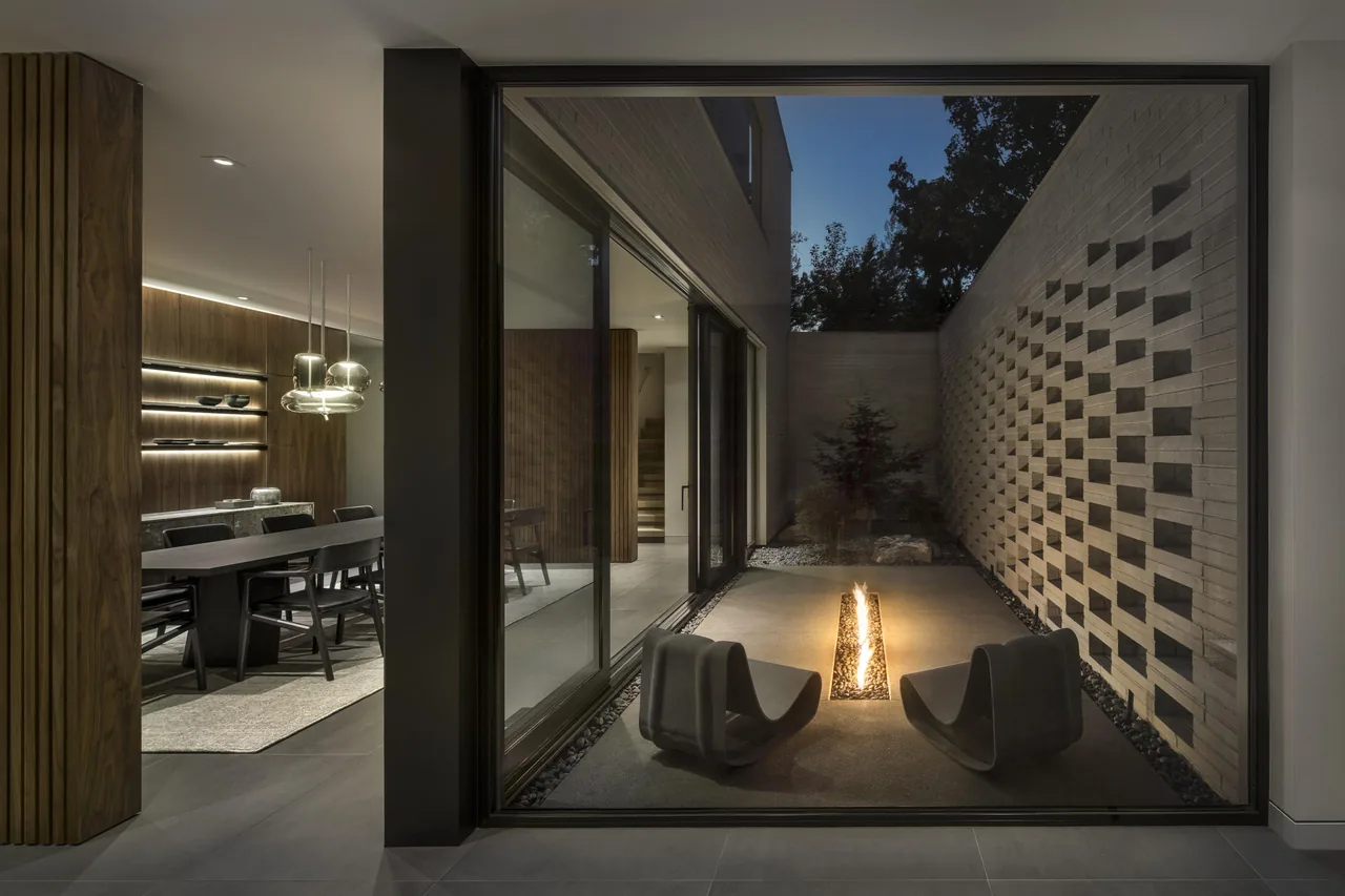 View of a nighttime courtyard with fire feature and sculptural seating visible from the dining room through large sliding glass doors.