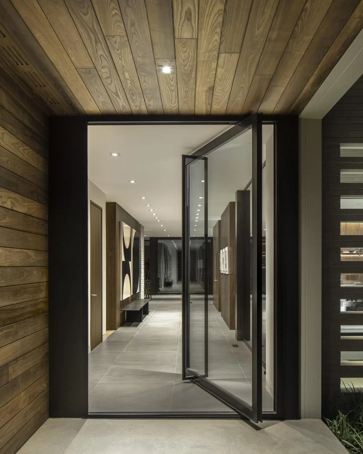 Interior hallway seen through a pivot glass door with wood paneling and recessed ceiling lights.