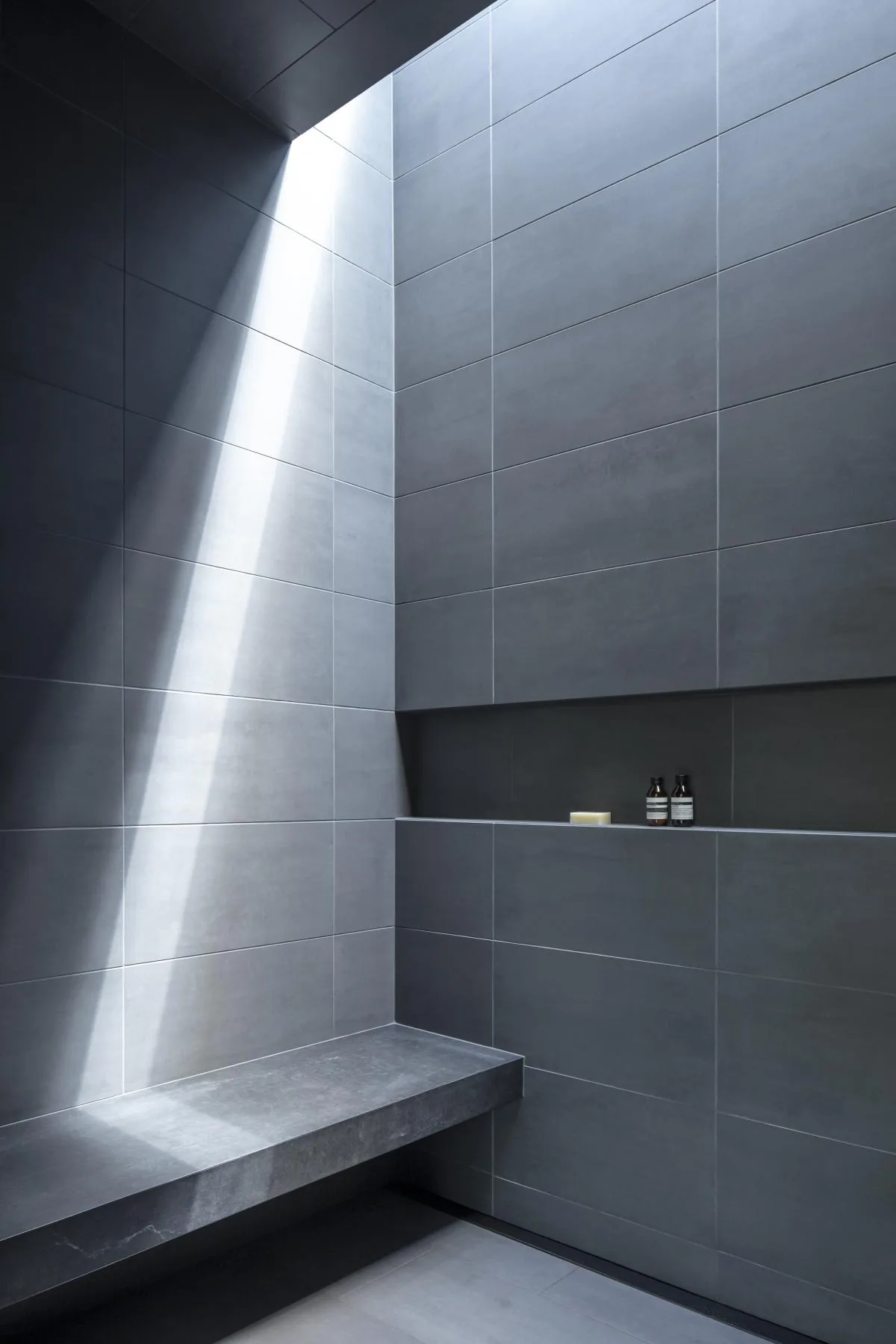 A minimalist shower features large gray tiles with a built-in shelf and natural light from a skylight above.