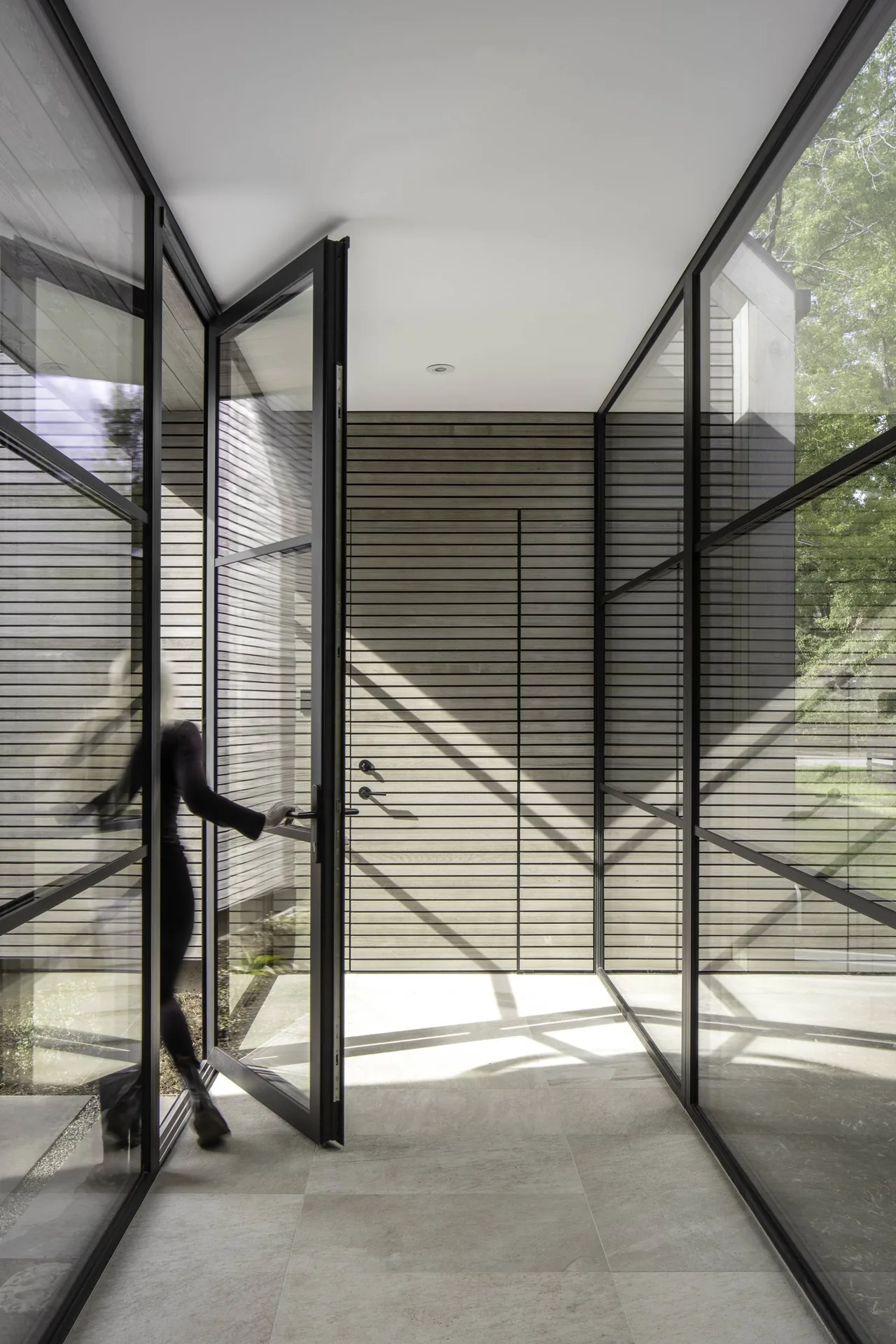 Entryway with large black-framed glass pivot door and horizontal wood siding wall at Linden Grove project.