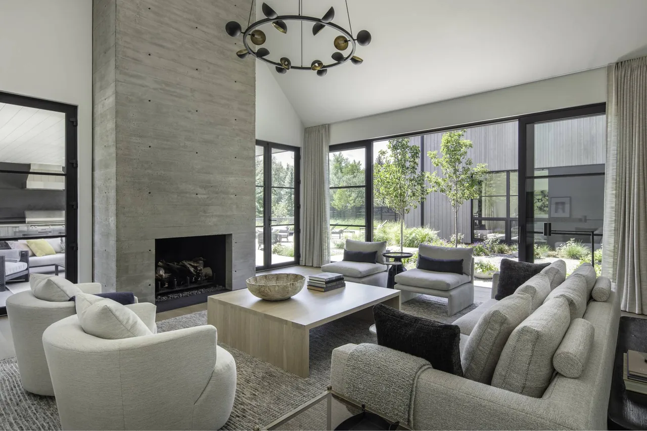 Living room features a tall exposed concrete fireplace wall, black window frames, and expansive glass walls overlooking a landscaped courtyard.