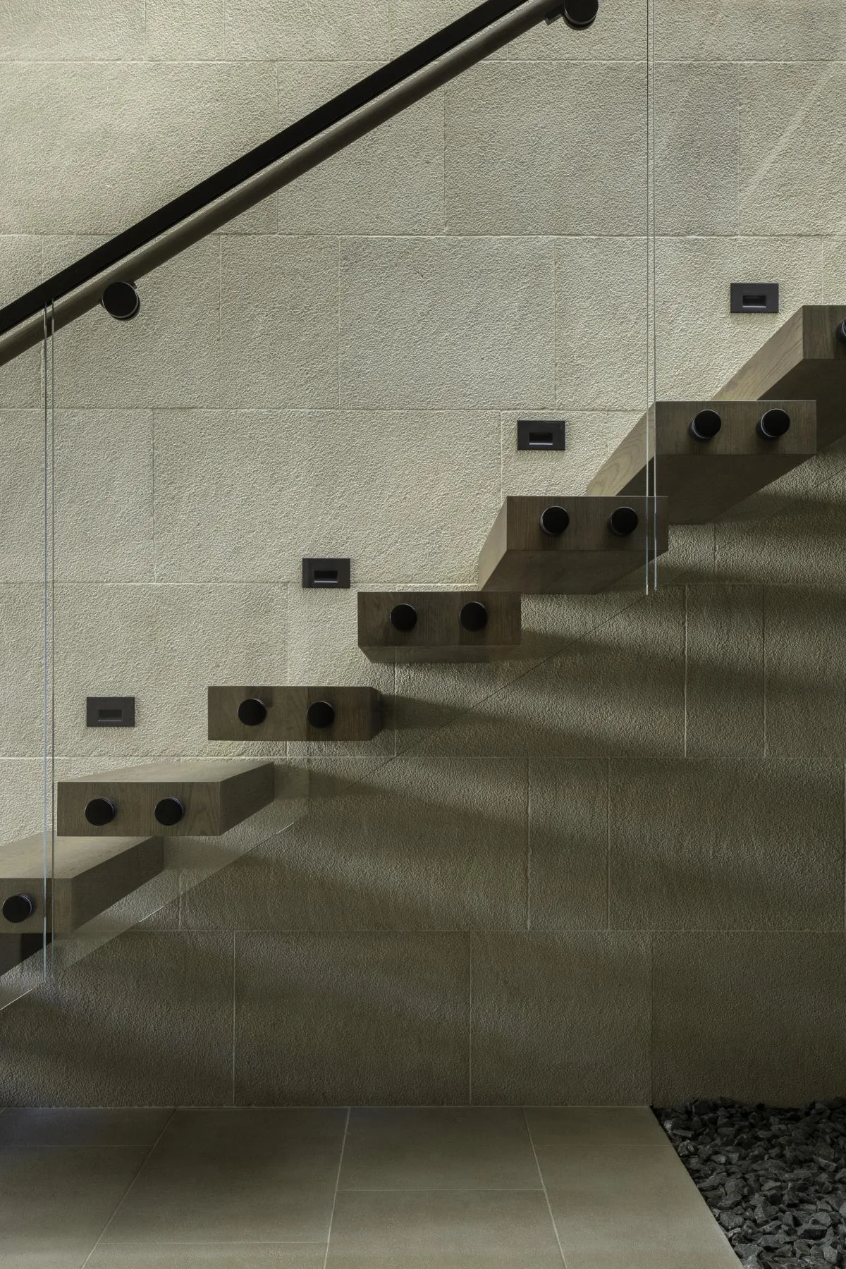Floating wooden staircase with glass balustrade attached to a textured stone wall.