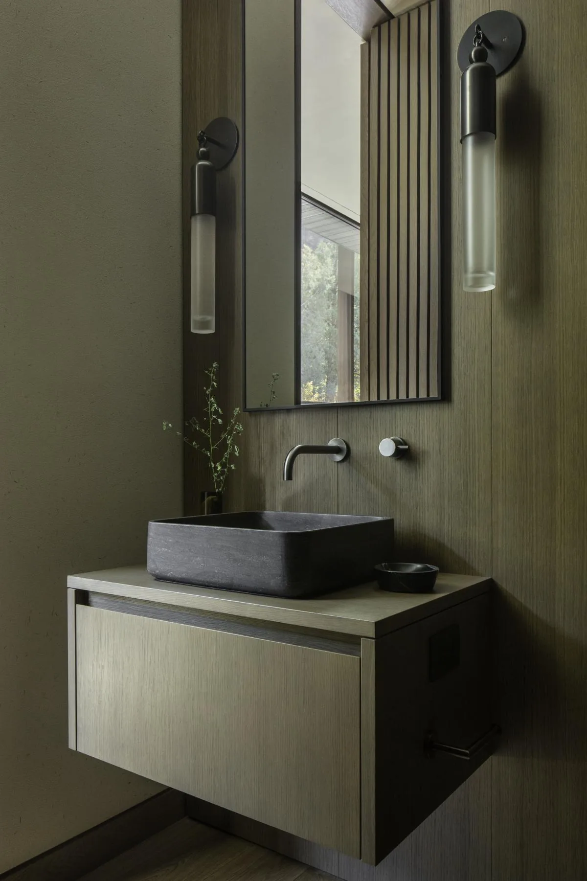 Bathroom vanity with black stone vessel sink under a vertical mirror flanked by wall sconces.