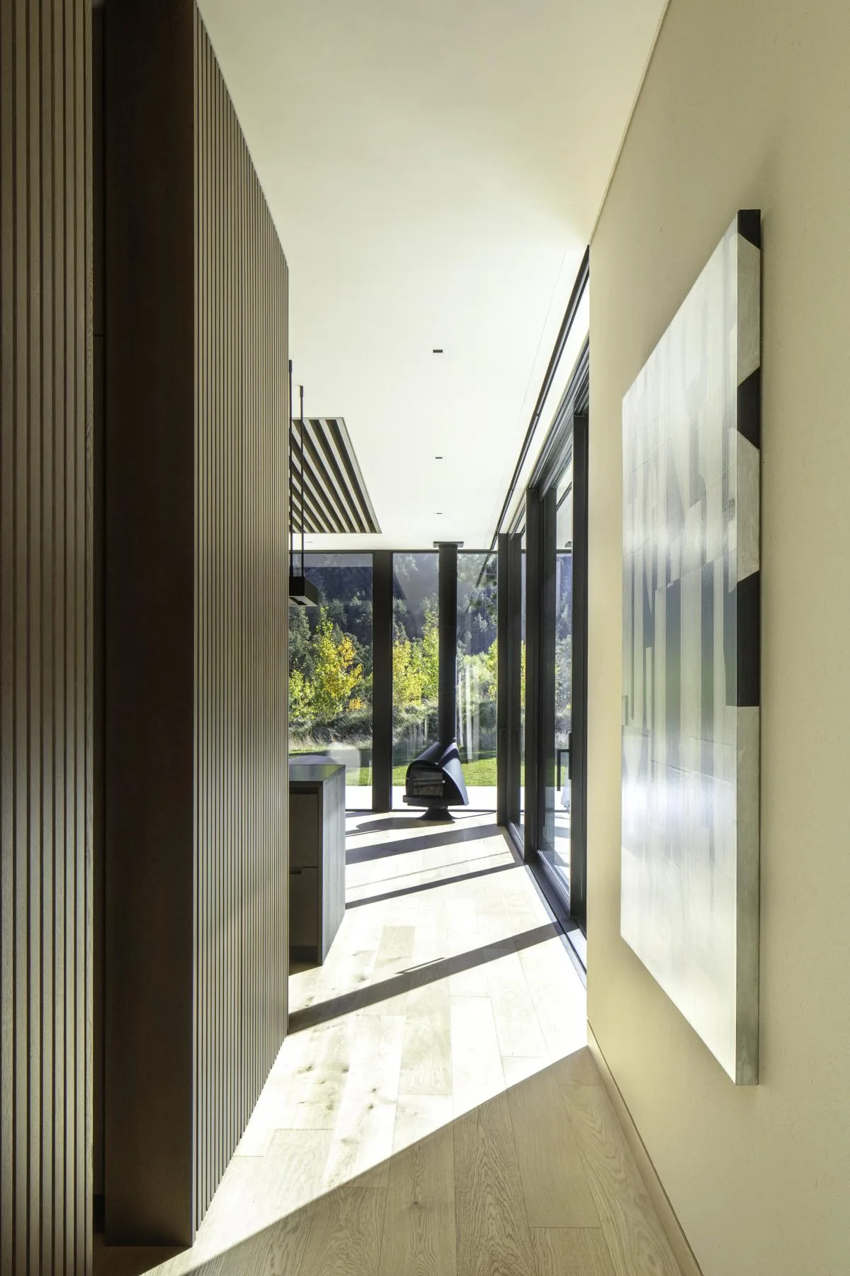 Interior view of a hallway with floor-to-ceiling glazing and wood paneling, showing sunlight patterns on the wooden floor.
