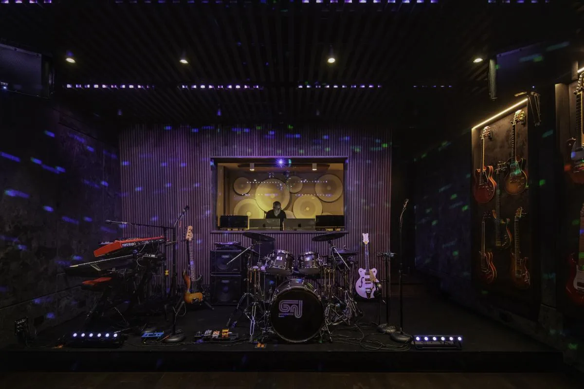 Music rehearsal room equipped with drums, multiple guitars, keyboards, and a control booth with soundproofing elements.