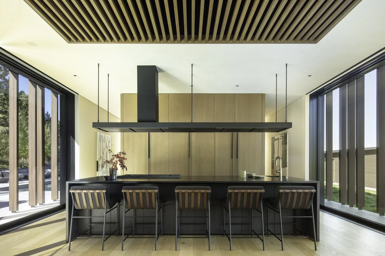 Interior view of a modern kitchen featuring a large island with a suspended black range hood and wood-paneled cabinetry.