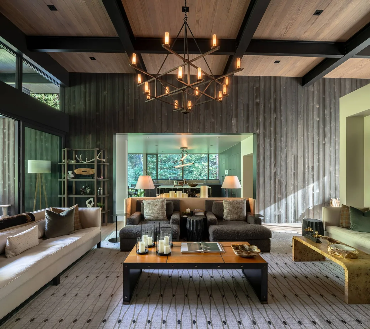 Living room features contrasting dark and light sofas, wood-paneled walls, and oversized chandelier with nature views outside.