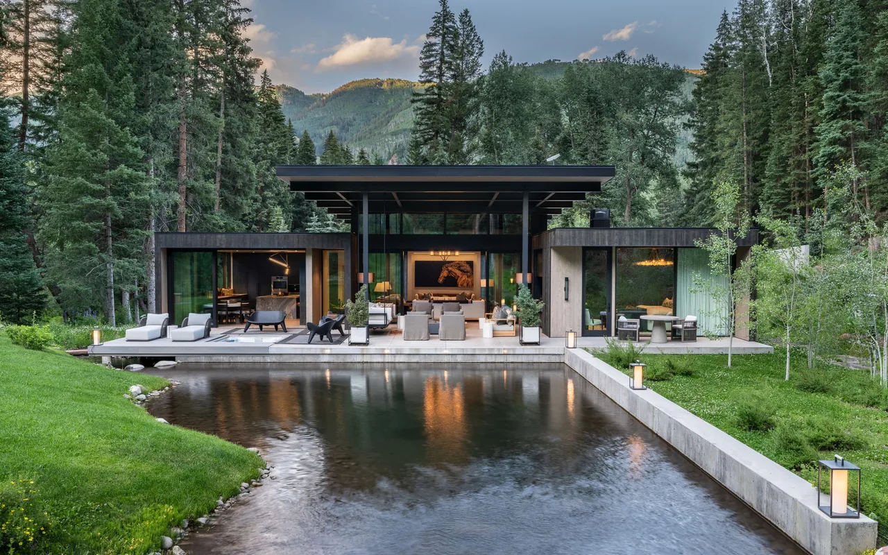 Modern flat-roof house with floor-to-ceiling glass walls overlooking a reflective pond surrounded by trees and mountains.