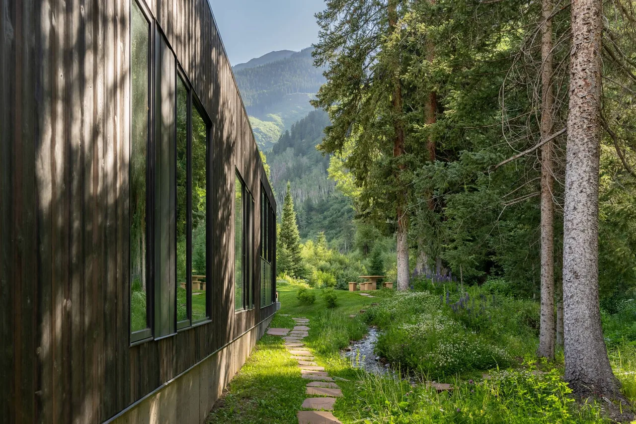 The Conundrum Creek house by Studio B Architects features wood siding with large vertical windows facing a creek and forested mountains.