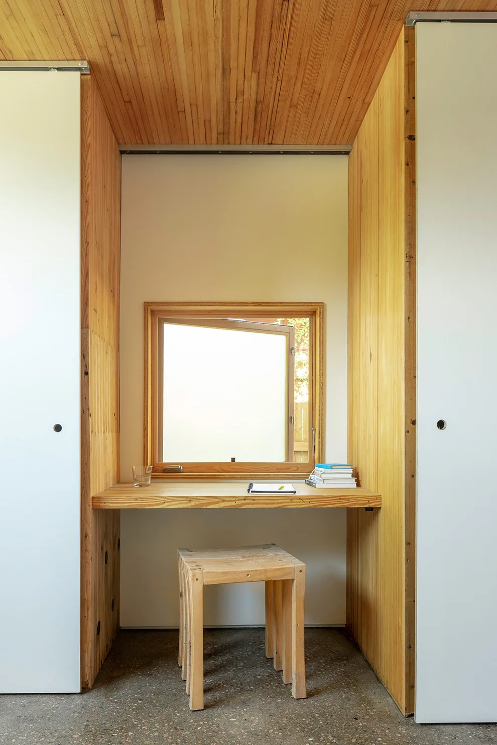 A compact wooden desk and stool set below a small square window in a wood-paneled nook with sliding white doors.