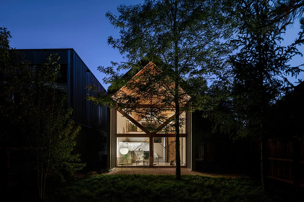 The A.D.U. project by Tres Birds Workshop shows a modern small house with illuminated interiors and exposed wooden structural features at night.
